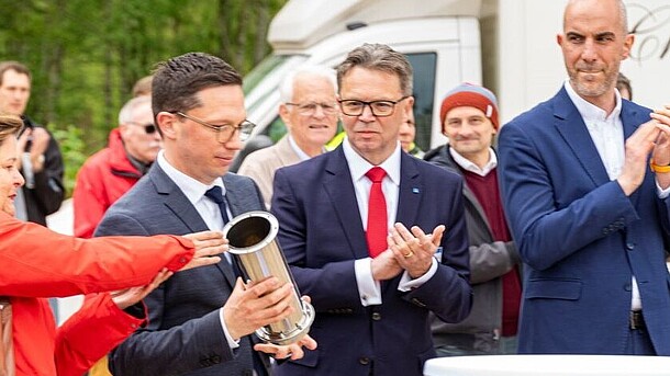 Three men in blue-grey suits stand outside, surrounded by men and women in casual wear. One man in the centre holds a silver pipe.