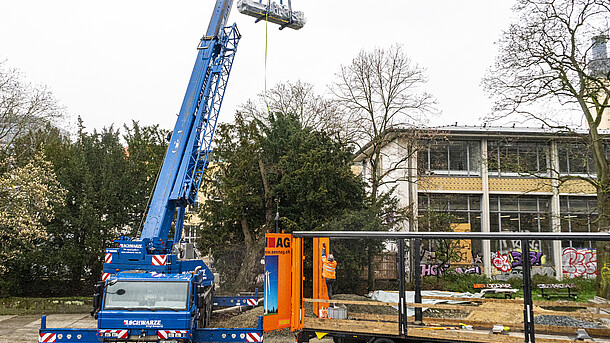 A large blue crane lifts a machine protected by transparent film over trees towards a yellowish-white building. In the front right of the picture stands a truck with the truck tarpaulins pushed to the side, on which the machine had previously been standing.