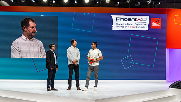  Three men stand on a large white stage and talk to each other.