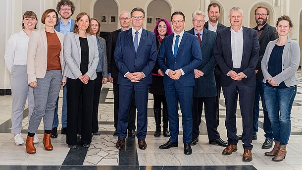 Sechs Frauen und neun Männer in Businesskleidung stehen in zwei Reihen neben- und hintereinander.